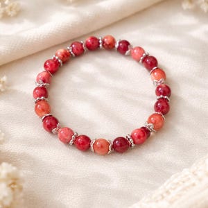Puede incluir: Un brazalete de cuentas con cuentas de piedras preciosas rojas y naranjas. El brazalete tiene detalles plateados entre las cuentas. Se muestra sobre una superficie blanca texturizada, con detalles florales en el fondo. Una joya.