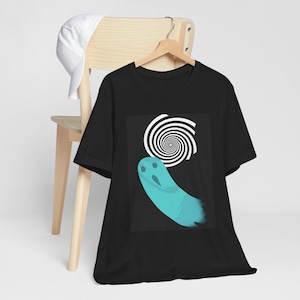 Peut inclure: T-shirt noir avec un motif de fantôme turquoise et un motif en spirale blanc. Le fantôme a trois trous pour les yeux et une queue flottante. Le t-shirt est suspendu à un cintre en bois.
