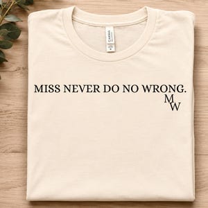 May include: A cream-coloured short-sleeve t-shirt with a crew neckline. The front features the black text MISS NEVER DO NO WRONG. followed by the letters M W stacked vertically in the bottom right corner of the text area. A white brand label is visible.