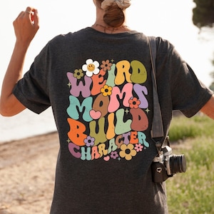 May include: A dark grey t-shirt with a colourful graphic on the back that says "Weird Moms Build Character" with flowers and a smiley face.