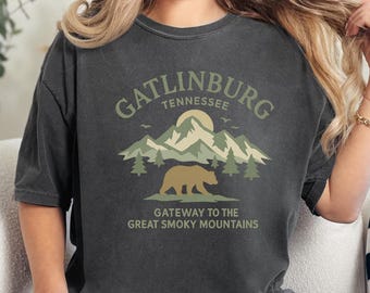 以下が含まれることがあります： ガトリンバーグ、テネシー州のグラフィックがプリントされた濃いグレーのTシャツ。デザインは山、木、クマ、そして「Gateway to the Great Smoky Mountains」の文字が特徴です。このTシャツはヴィンテージ風です。