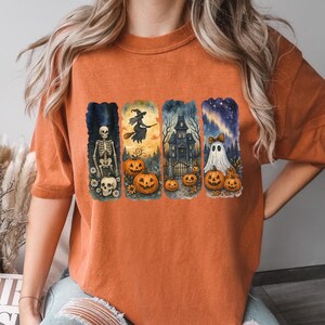 May include: Orange short-sleeved t-shirt featuring a Halloween-themed graphic. The graphic includes a skeleton, witch, haunted house, ghost, pumpkins, and a starry night sky. The design is in a watercolor style.