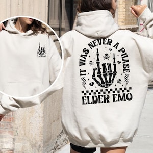 May include: A beige hoodie with a black graphic on the back that says "IT WAS NEVER A PHASE" with a skeleton hand, skulls, and lightning bolts. The front of the hoodie has a smaller graphic of a skeleton hand and the words "ELDER EMO".