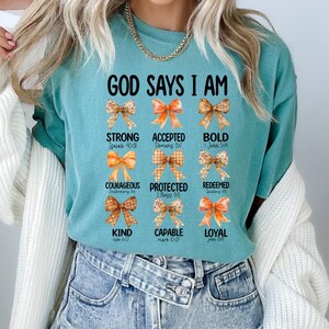 Puede incluir: Camiseta turquesa con la frase "GOD SAYS I AM" y palabras como "STRONG" y "BOLD" con referencias bíblicas. El diseño incluye lazos decorativos en tonos naranja, marrón y amarillo.
