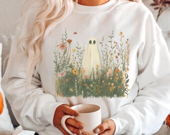 May include: White sweatshirt featuring a whimsical ghost design amidst a field of wildflowers. The ghost has a simple, friendly face. The floral elements include various colors, creating a soft, natural aesthetic. A cozy, seasonal apparel item.