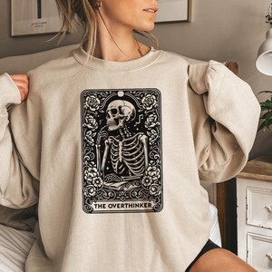 May include: A beige sweatshirt with a black and white graphic of a skeleton within a decorative frame, adorned with floral details. The text "THE OVERTHINKER" is displayed at the bottom. The sweatshirt is a neutral tone and has long sleeves.