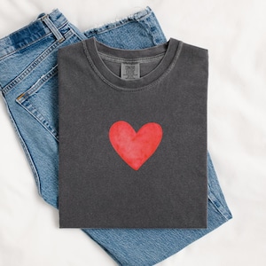 Peut inclure: Un t-shirt gris anthracite avec un motif de cœur rouge. Il est associé à un jean bleu clair, créant un look décontracté. Le t-shirt a un col rond et des manches courtes, idéal pour un style quotidien.