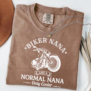 以下が含まれることがあります： 「BIKER NANA」の文字と白いバイクのグラフィックがプリントされた茶色のTシャツ。「LIKE A NORMAL NANA - Only Cooler」の文字も含まれています。ペンダント付きのシルバーのネックレスがTシャツの上にあります。