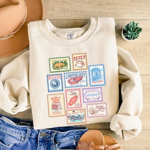May include: Cream-colored sweatshirt featuring a collection of colorful postage stamp designs. The stamps depict various images, including a bear, coffee cup, lobster, Ferris wheel, and books. The sweatshirt is paired with blue denim jeans and a brown hat.