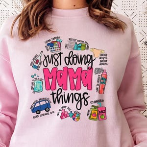 May include: Pink sweatshirt with a graphic design that says "Just doing mama things" and features a variety of illustrations of items associated with motherhood, such as a car seat, diapers, and a bottle of hand sanitizer.