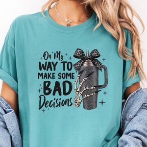 Könnte beinhalten: Tealblaues T-Shirt mit einer schwarzen Grafik einer Skeletthand, die einen Glitzerbecher mit einer schwarzen Schleife hält. Der Text auf dem Shirt lautet "On my way to make some bad decisions".