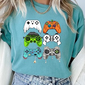 Op de afbeelding: Tealblauw T-shirt met een afbeelding van vijf verschillende gekleurde videogamecontrollers met Halloween-thema's. De controllers zijn een geest, een pompoen, Frankenstein, een skelet en een mummie.