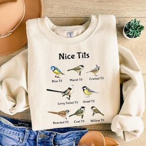 May include: A cream-colored sweatshirt with the text "Nice Tits" printed on the front. The text is surrounded by illustrations of different types of birds, each labeled with its name, such as "Blue Tit", "Marsh Tit", and "Crested Tit".
