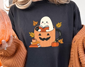 Op de afbeelding: Zwart T-shirt met een afbeelding van een geest die een boek leest in een pompoenmok. De geest heeft een schattige glimlach en houdt een boek vast. De mok is omringd door herfstbladeren en een stapel boeken.