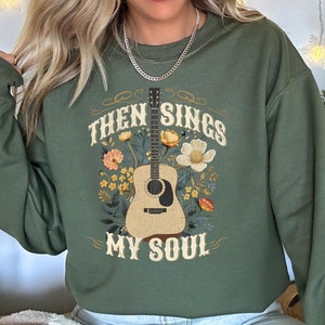 May include: Olive green sweatshirt featuring a guitar graphic with floral accents and the text "Then Sings My Soul." The design includes a detailed acoustic guitar and various flowers in shades of yellow, orange, and white.