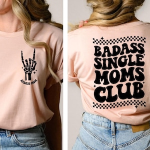 May include: A light pink t-shirt with a black skeleton hand making a rock and roll sign on the front and the text "Badass Mom" underneath. The back of the shirt has the text "Badass Single Moms Club" in black with a checkered pattern.