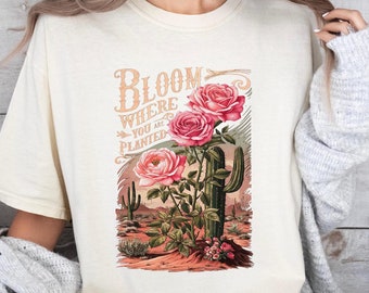 May include: A cream colored t-shirt with a graphic print of pink roses and a cactus. The text "Bloom Where You Are Planted" is printed on the shirt.