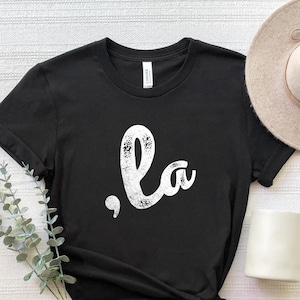 May include: Black t-shirt with a white distressed graphic that says ",la" on it. The shirt is laid out on a white surface with a hat and a mug.