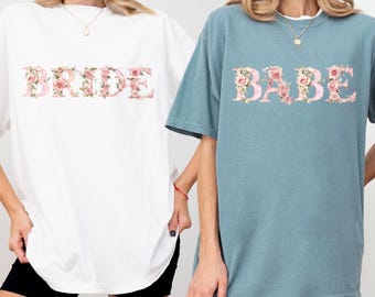May include: Two oversized t-shirts, one white and one ice blue. The white shirt says "BRIDE" in floral letters. The ice blue shirt says "BABE" in floral letters. Both shirts are short-sleeved.