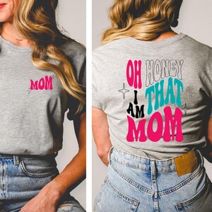 Puede incluir: Una camiseta gris con la palabra "MOM" en rosa en la parte delantera y el texto "OH HONEY I AM THAT MOM" en rosa, negro y turquesa en la parte trasera.