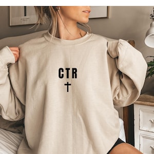 Op de afbeelding: Beige sweatshirt met de zwarte tekst "CTR" boven een kruis. De top met lange mouwen heeft een ronde hals en een relaxte pasvorm. De afbeelding toont een close-up van de sweatshirt.