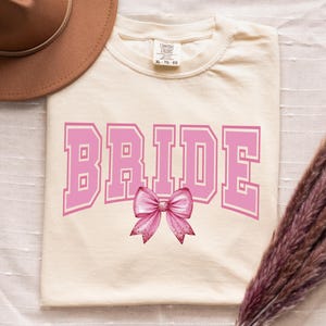 May include: A cream colored t-shirt with the word "BRIDE" in pink letters with a pink bow underneath.