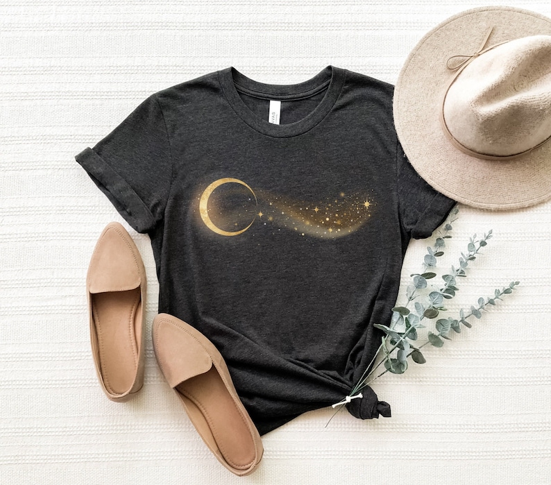 May include: A dark grey t-shirt with a gold crescent moon and a trail of gold stars printed on the front. The shirt is laid flat on a white surface with a pair of beige shoes and a brown hat.