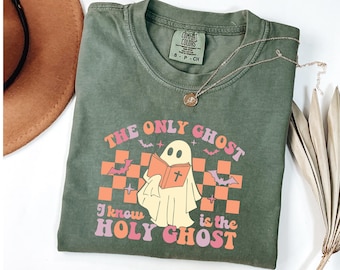 Op de afbeelding: Olijfgroen t-shirt met de tekst "The Only Ghost I Know is the Holy Ghost" in een retro design. De afbeelding toont een spook dat een boek leest, vleermuizen en een dambordpatroon. Een gouden ketting ligt op het t-shirt.