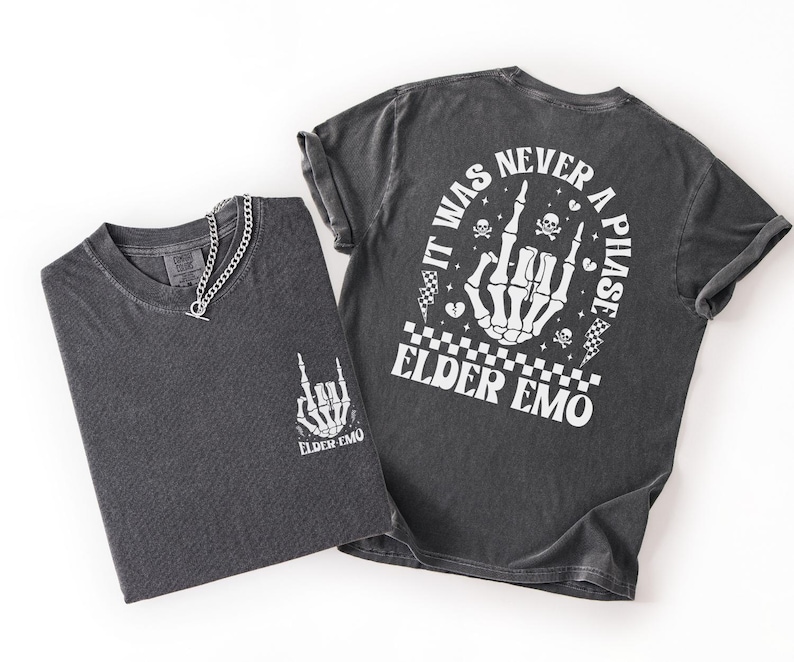 K&ouml;nnte beinhalten: Zwei anthrazitfarbene T-Shirts mit wei&szlig;en Grafikdesigns. Ein Shirt zeigt eine Skeletthand mit dem Text "ELDER EMO" und eine Halskette. Das andere Shirt hat den Text "IT WAS NEVER A PHASE" um eine Skeletthand, Totenk&ouml;pfe und Schachbrettflaggen.
