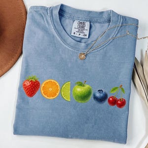 May include: A light blue Comfort Colors t-shirt featuring a colorful fruit design. The design includes a strawberry, orange slice, lime wedge, green apple, blueberry, and cherries. A gold necklace is draped across the shirt.