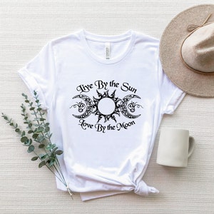May include: White t-shirt with a black graphic of a sun and moon with the text "Live By the Sun Love By the Moon".
