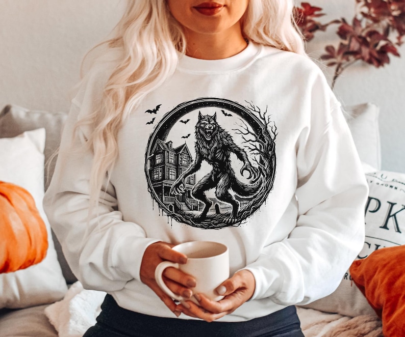 May include: White sweatshirt featuring a black and white illustration of a werewolf in a circular frame. The design includes a haunted house, bare trees, and bats, creating a spooky Halloween theme. The sweatshirt is a comfortable, casual style.