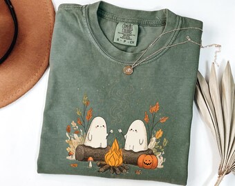 Può includere: T-shirt Comfort Colors verde salvia con una scena di falò con due personaggi fantasma che arrostiscono marshmallow. Il design include una zucca, foglie d'autunno e un tronco. Una collana d'argento con un ciondolo è appoggiata sulla maglietta.