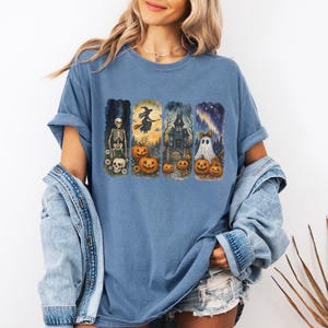 May include: A blue t-shirt featuring a Halloween-themed design. The graphic includes a skeleton, witch, haunted house, and ghost, each with pumpkins. The shirt has short sleeves and a relaxed fit.