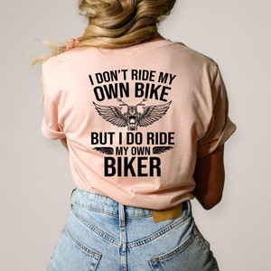 以下が含まれることがあります： 「I DON'T RIDE MY OWN BIKE BUT I DO RIDE MY OWN BIKER」の黒い文字がプリントされた、薄いピンク色のTシャツ。翼のついたバイクのグラフィックがテキストの上に配置されています。ブロンドの髪の人が着ています。