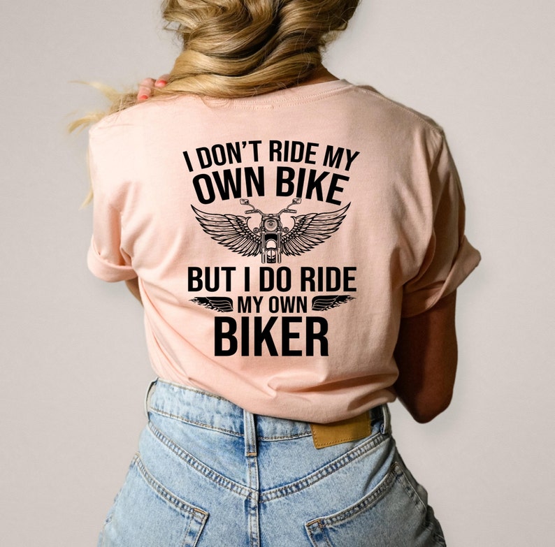 以下が含まれることがあります： 「I DON'T RIDE MY OWN BIKE BUT I DO RIDE MY OWN BIKER」の黒い文字がプリントされた、薄いピンク色のTシャツ。翼のついたバイクのグラフィックがテキストの上に配置されています。ブロンドの髪の人が着ています。