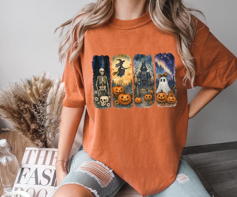 May include: Orange short-sleeved t-shirt featuring a Halloween-themed graphic. The graphic includes a skeleton, witch, haunted house, ghost, pumpkins, and a starry night sky. The design is in a watercolor style.