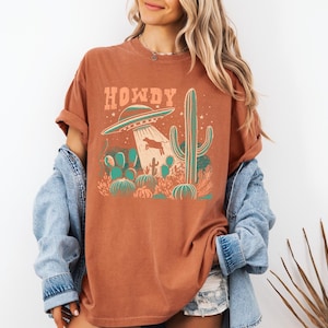 May include: Rust-colored t-shirt with a graphic featuring a UFO beaming up a cow, cacti, and the word "HOWDY" in orange. The design has a vintage, desert-themed aesthetic.