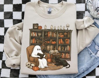 Puede incluir: Sudadera beige con una ilustración caprichosa de un fantasma leyendo en una biblioteca acogedora. El diseño incluye una estantería, calabazas, velas, un gato negro y calaveras, ideal para Halloween o amantes de los libros.