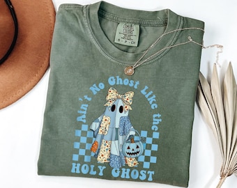 Op de afbeelding: Olijfgroen T-shirt met een afbeelding van een spook met een pompoenemmer. Het spook draagt een patchwork outfit en een strik. De tekst op het shirt luidt "Ain't No Ghost Like the Holy Ghost". Een gouden ketting ligt over het shirt.