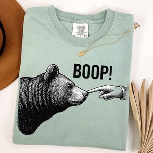 May include: A light green t-shirt with a graphic of a bear's head being touched on the nose by a hand, with the word "BOOP!" in black. A gold necklace and a brown hat are also visible.