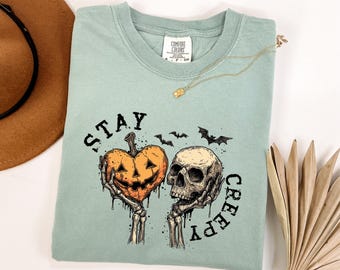 May include: A pale green Comfort Colors t-shirt with a Halloween design. The graphic shows a heart-shaped pumpkin and a skull held by skeleton hands, with the words "STAY CREEPY" in black.