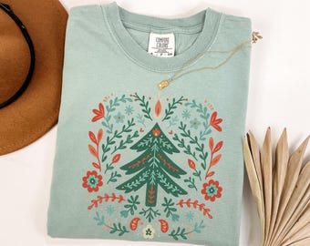 Puede incluir: Una camiseta verde claro con un diseño de árbol de Navidad. El árbol está rodeado por una corona de hojas y flores en rojo, verde y turquesa. Un collar dorado está sobre la camiseta. Un sombrero marrón es visible en la esquina superior izquierda.