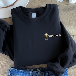 May include: Black sweatshirt with the text "VITAMIN G" and a graphic of a pint of beer. The sweatshirt is folded on top of a pair of blue jeans and a white hat.