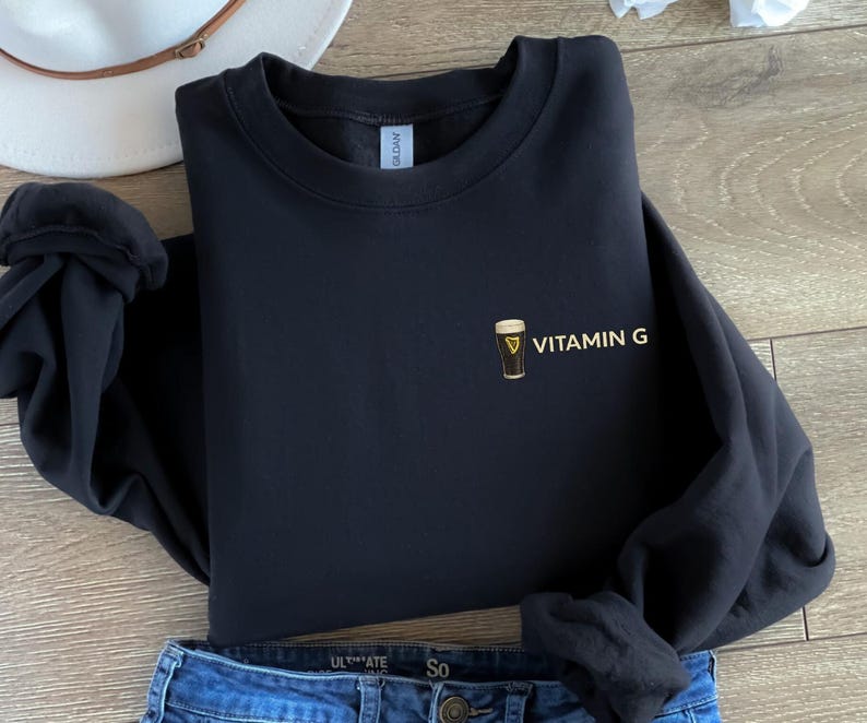 May include: Black sweatshirt with the text "VITAMIN G" and a graphic of a pint of beer. The sweatshirt is folded on top of a pair of blue jeans and a white hat.