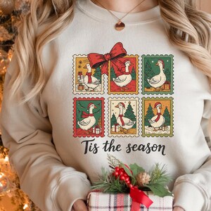 Puede incluir: Sudadera color crema con un diseño de seis sellos postales con gansos de dibujos animados con gorros y bufandas de Papá Noel. Un gran lazo rojo está encima de los sellos, con el texto "Tis the season" debajo.