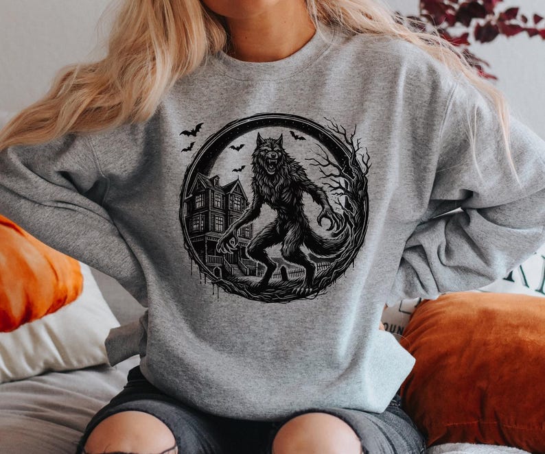 May include: A gray sweatshirt featuring a black and white illustration of a werewolf standing in front of a haunted house. The design is framed by a circular border with bats and bare tree branches, perfect for Halloween.
