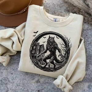 May include: Cream-colored sweatshirt featuring a black and white werewolf graphic inside a circular frame. The design includes a haunted house, bats, and bare trees, creating a spooky Halloween theme. The sweatshirt has a crew neck and long sleeves.