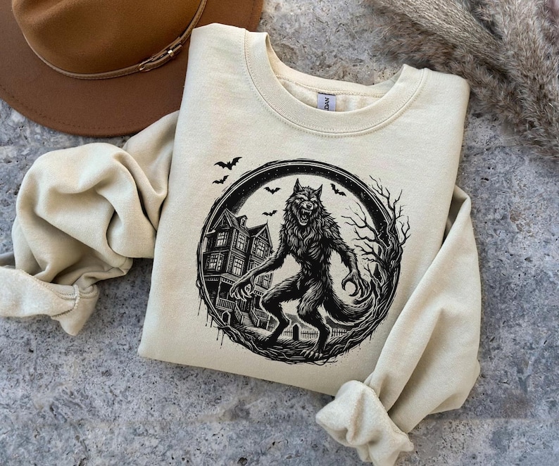 May include: Cream-colored sweatshirt featuring a black and white werewolf graphic inside a circular frame. The design includes a haunted house, bats, and bare trees, creating a spooky Halloween theme. The sweatshirt has a crew neck and long sleeves.