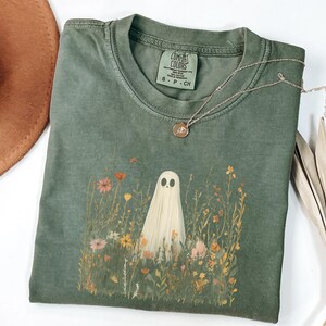 May include: Sage green t-shirt with a ghost graphic in a field of wildflowers. The shirt has a crew neck and short sleeves. A gold necklace with a small pendant rests on the shirt. A brown hat is visible in the upper left corner.